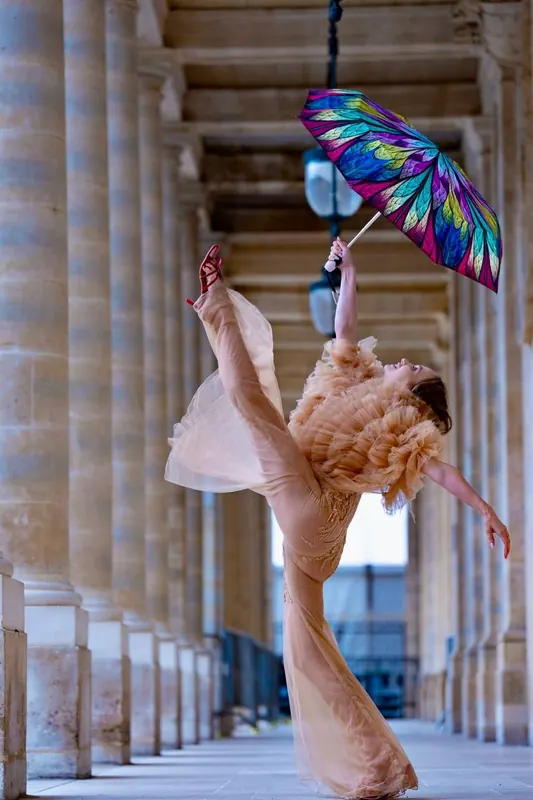Zuza in a long colonnade, holding a bright patterned umbrella high